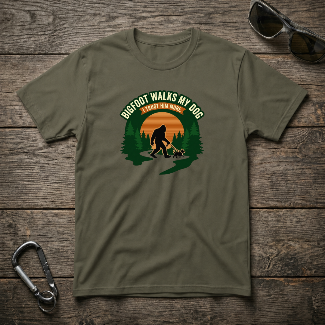 Military green Bigfoot walks a dog graphic T-shirt by Designed For The Wild Co, showcasing a playful outdoor scene.
