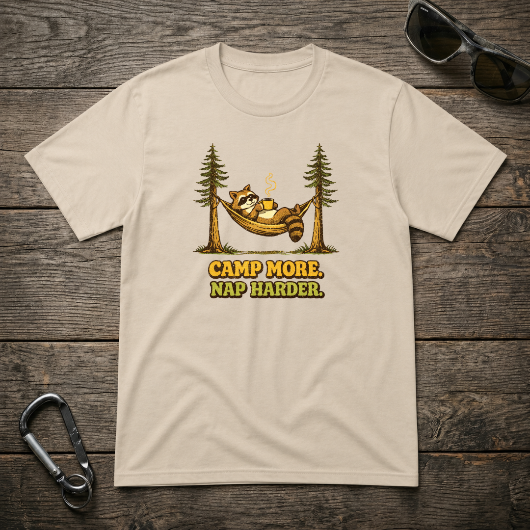 Graphic T-shirt from Designed For The Wild Co featuring "Camp More, Nap Harder" slogan, with a natural outdoor scene in earthy tones.